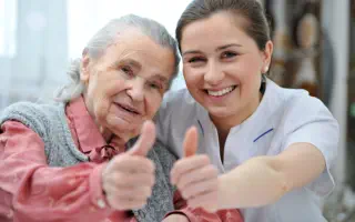 Pflegerin mit Patientin, beide lächeln und zeigen den Daumen nach oben.